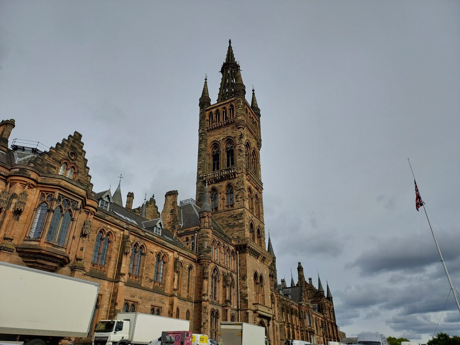 A photograph of the main building of the University of Glasgow. The sky is cloudy and the UK flag is visible.